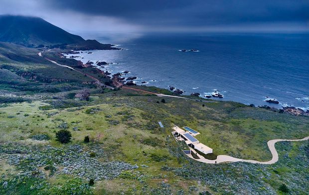 Aerial view of a low-profile modern residence set within expansive coastal terrain, with a winding driveway approaching the building. The property overlooks a rugged rocky shoreline and open ocean, with a mountainous headland and sparse natural vegetation surrounding the site. The architecture appears elongated and integrated into the landscape, with flat roof planes and strong geometric forms.