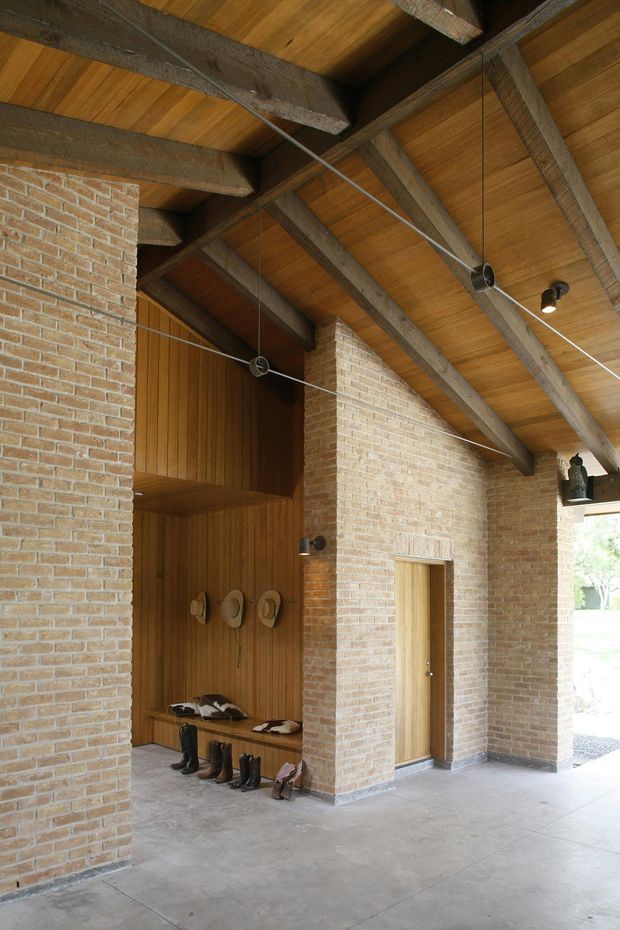 A covered entry or mudroom-like architectural space with exposed timber roof beams and timber soffit, pale brick walls, a concrete floor, and a recessed timber-clad alcove containing a bench, hooks, and footwear storage. A simple timber door is set into the brick wall, while suspended cable lighting and black spotlights provide illumination. The space opens to the outdoors at the right.