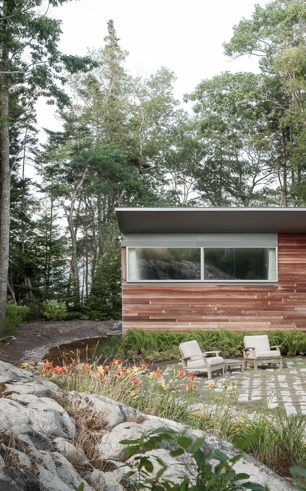 Exterior view of a low modern building set within a wooded landscape. The facade combines horizontal timber cladding with a broad strip window and a projecting flat roof, opening onto a landscaped stone-paved terrace with outdoor lounge chairs, ornamental grasses, flowering plants, and natural boulders.
