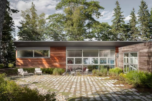 Single-storey contemporary home viewed from a landscaped courtyard. The flat-roofed facade combines warm horizontal timber cladding, extensive glazing, and a shingle-clad projecting volume. A paved courtyard with regular stone grid paving and planted borders occupies the foreground, with mature trees surrounding the property.