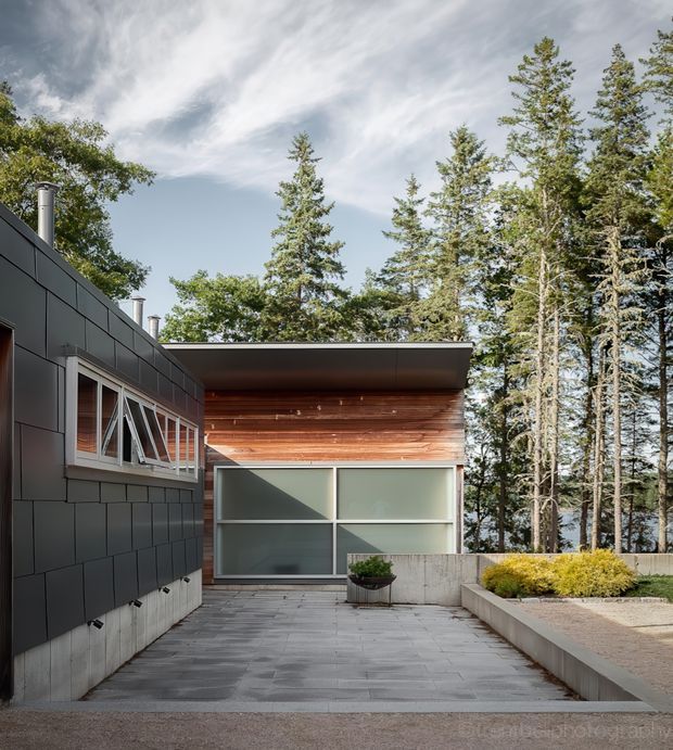 Exterior view of a contemporary, low-profile house in a wooded setting. The facade combines dark rectangular cladding with a concrete lower wall, horizontal timber cladding beneath a broad flat roof overhang, and large frosted glass garage or entry doors. A paved courtyard and raised landscaped planter occupy the foreground, with mature coniferous trees surrounding the building.