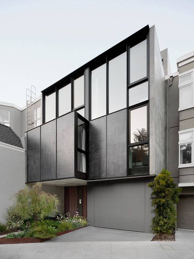 Street-facing exterior photograph of a narrow contemporary multi-storey townhouse or urban residence. The facade combines dark metal-looking panels, extensive black-framed glazing, a projecting upper volume, and a grey garage door, set among closely adjoining traditional neighbouring homes. A small landscaped front garden and driveway occupy the foreground.