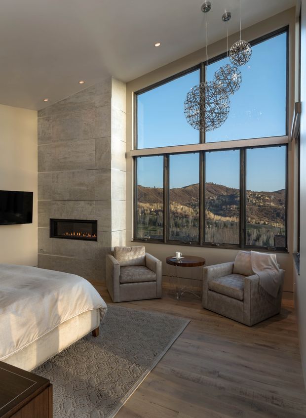 A refined bedroom with a partially visible upholstered bed, two plush armchairs, and a small round side table arranged beside expansive double-height dark-framed windows. A tall concrete-look feature wall incorporates a linear fireplace and wall-mounted television. Sculptural spherical pendant lights hang in front of the glazing, which overlooks a dry mountainous landscape. The room has warm timber flooring, a large patterned rug, neutral upholstery, and soft natural light.
