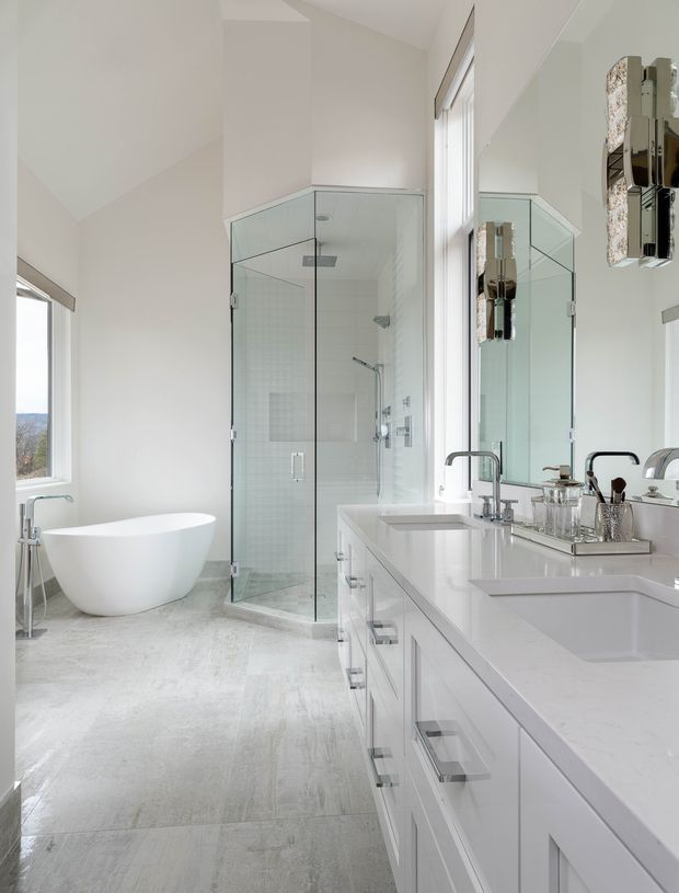 Bright, spacious contemporary bathroom with a freestanding white bathtub, large frameless glass shower enclosure, double vanity with white cabinetry and integrated basins, wall-to-wall mirror, chrome fixtures, and pale stone-look flooring. The room has abundant natural light and a clean, refined aesthetic.