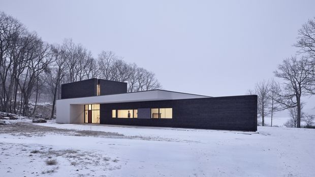 A minimalist single-storey modern house set in a snowy wooded landscape. The low, elongated form combines a dark horizontal masonry or cladding wall with a crisp white rendered volume, a flat roof, and a raised dark rectangular rooftop volume. Narrow horizontal windows and a glazed entry punctuate the facade.