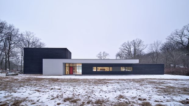 A minimalist contemporary single-storey residence viewed from the front across a snow-covered landscape. The building has a strong horizontal composition, dark charcoal cladding, smooth white rendered walls, a projecting flat roof, and long bands of glazing. A taller dark rectangular volume rises at the rear, while leafless trees form the background.