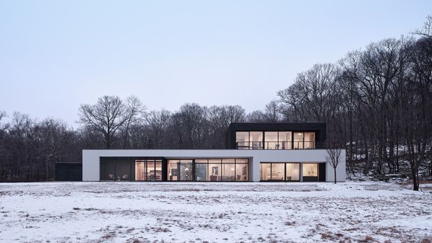 A minimalist two-storey modern house set in a snowy, wooded landscape. The building has a low horizontal ground floor with extensive floor-to-ceiling glazing and a dark upper-level rectangular volume projecting above it. The facade combines smooth light-coloured wall surfaces, dark cladding or trim, and large illuminated windows. Bare deciduous trees and snow-covered open ground surround the residence.