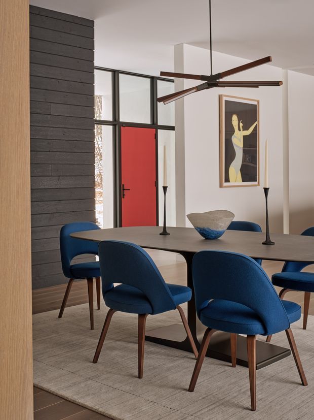 A refined contemporary dining room with a long dark dining table surrounded by upholstered navy-blue dining chairs with timber legs. A large sculptural ceiling fan hangs above, while a black horizontal timber feature wall, black-framed glazing, and a vivid red entry door create strong architectural accents. Neutral walls, a textured pale rug, minimalist candleholders, and framed abstract artwork complete the space.