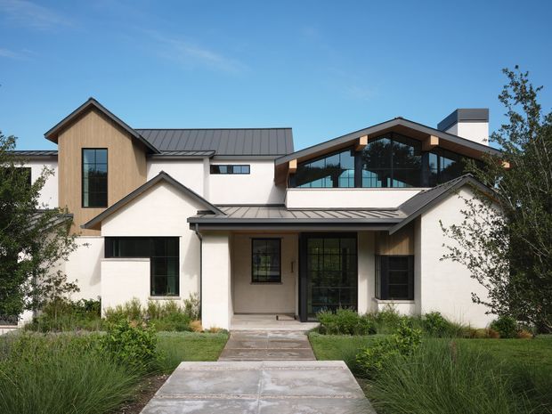 Front exterior view of a large contemporary residence with multiple intersecting pitched roof forms, dark standing-seam metal roofing, white rendered walls, warm timber-clad upper volumes, and extensive black-framed glazing. A central paved path leads through a landscaped garden to the recessed entry. The house has a strong gabled composition and a prominent glazed upper-level living volume.