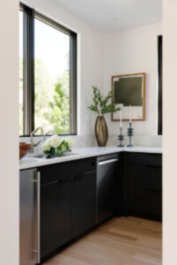 A bright contemporary kitchen corner with an L-shaped run of matte black cabinetry, pale stone benchtops, integrated stainless-steel dishwasher, black-framed window, timber-look flooring, and a simple decorative arrangement of a vase, framed artwork, and candlesticks. Natural light enters through the large window above the sink.