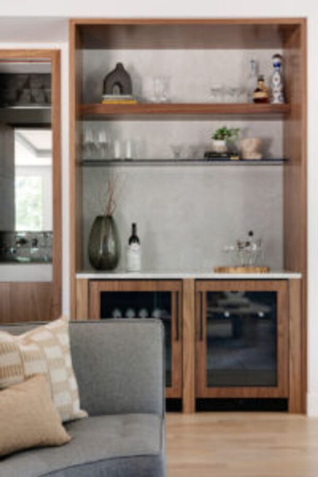 A built-in timber display and bar cabinet with open shelving, glass shelves, decorative objects, wine bottles and glassware. The lower cabinetry has dark glazed doors, while a grey upholstered sofa occupies the foreground. Warm timber tones, pale walls and light timber flooring create a refined residential living area.