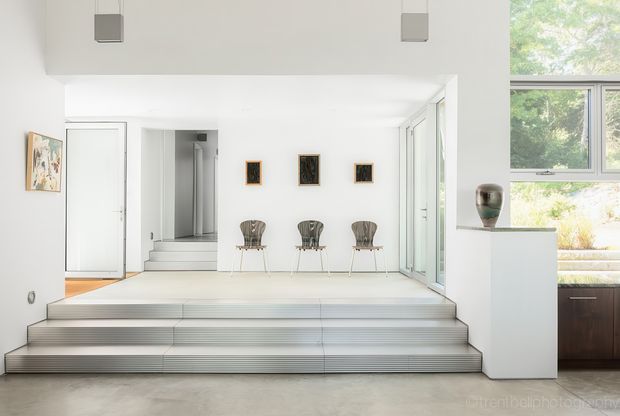 Bright minimalist interior with a broad set of shallow steps leading to a raised white gallery-like living area. The space has white walls and ceilings, pale flooring, full-height glazed doors, a stairway to an upper level, three sculptural chairs, framed artwork, and large windows overlooking greenery. A white pedestal and small sculpture sit at right, with part of a dark timber kitchen or joinery area visible beyond.