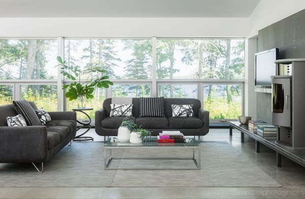 Bright contemporary living room with floor-to-ceiling windows overlooking trees, a dark charcoal sectional and matching sofa, glass coffee table, indoor plants, and a wall-mounted television with built-in media joinery. The room has a spacious, airy arrangement and a restrained neutral palette.