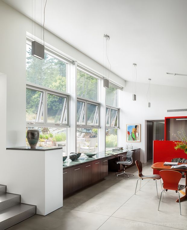 Bright, high-ceilinged contemporary interior with a long wall of large operable windows overlooking greenery. Dark wood built-in cabinetry and a work desk run beneath the glazing, while sculptural pendant lights hang from the white ceiling. A vivid red built-in partition or joinery element defines the dining area, furnished with modern red and orange chairs around a round table. Steps with dark striped edging lead into the space, and artwork and decorative objects add colour.