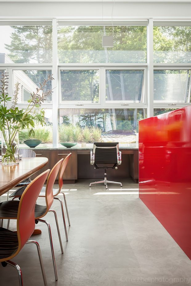 Bright contemporary open-plan interior combining a dining area and compact home office. Floor-to-ceiling white-framed glazing overlooks a leafy garden, while a long timber dining table with curved red chairs occupies the left side. A black office chair sits at a built-in timber desk beneath the windows, and a vivid glossy red full-height partition or cabinet forms a strong feature on the right. The space has polished concrete flooring, abundant natural light, and clean modern lines.