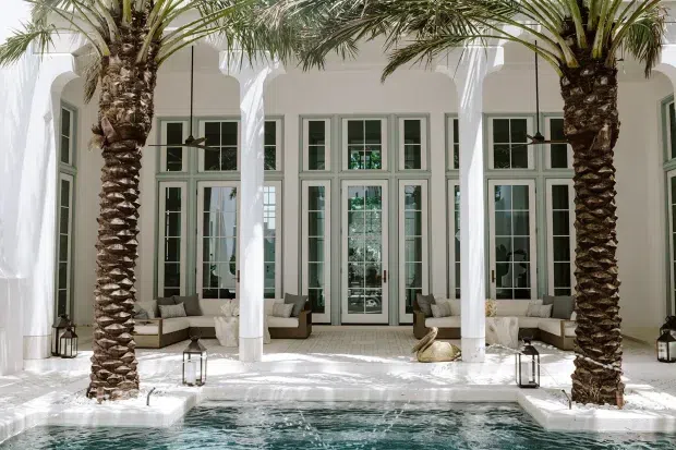 A bright, resort-style courtyard pool framed by two mature palm trees, white stucco walls, tall arched openings, slender white columns, and full-height multi-pane French doors. The covered terrace contains built-in or upholstered lounge seating, lanterns, and subtle decorative accents, creating a luxurious Mediterranean-inspired outdoor living space.