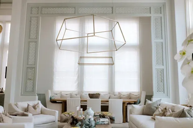 Elegant, bright living and dining space with a large bay-style window framed by intricate decorative lattice panelling. A geometric brass pendant hangs above a dining table with upholstered white chairs and built-in window seating, while white sofas and armchairs create a refined conversational area in the foreground. The palette is predominantly soft white and cream with warm metallic accents.