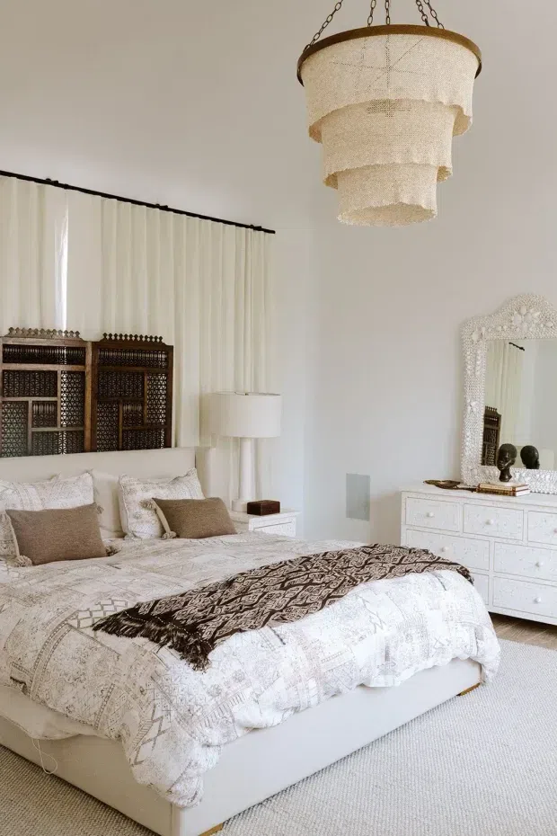 Bright, refined bedroom with a low upholstered bed dressed in layered white and taupe patterned bedding, an ornate dark carved screen-style headboard, white bedside table, and a white dresser topped with an elaborate textured mirror. A tiered woven pendant light hangs from the ceiling, while sheer curtains soften the window wall. The palette is predominantly white, cream, beige, and brown with an eclectic global-inspired decorative feel.