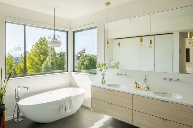 Bright contemporary bathroom with a freestanding white bathtub positioned beside large corner windows, and a long light-wood double vanity with integrated basins beneath mirrored cabinetry. Minimal brass pendant lights hang above the vanity, while grey floor tiles and pale walls create a clean, airy setting with leafy views outside.