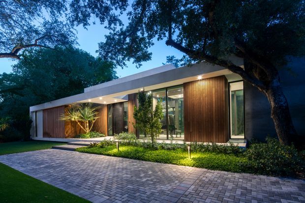 A single-storey contemporary house photographed at dusk, with a long low flat roof, extensive floor-to-ceiling glazing, warm vertical timber wall cladding, and white rendered or painted facade elements. The home is set among mature trees and lush landscaping, with a broad paved driveway or forecourt and subtle landscape lighting illuminating the garden and facade.