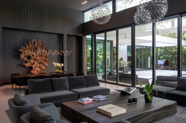 A sophisticated, spacious living room with floor-to-ceiling glass doors and windows opening toward a landscaped outdoor courtyard or terrace. The room features a large dark sectional sofa, a substantial dark wood or wood-look coffee table, a dramatic sculptural branch chandelier, two spherical pendant lights, dark vertically textured wall panelling, and a dining area with additional sculptural lighting. The palette is predominantly charcoal, black, dark brown, and muted green, creating a refined contemporary atmosphere with strong indoor-outdoor connection.