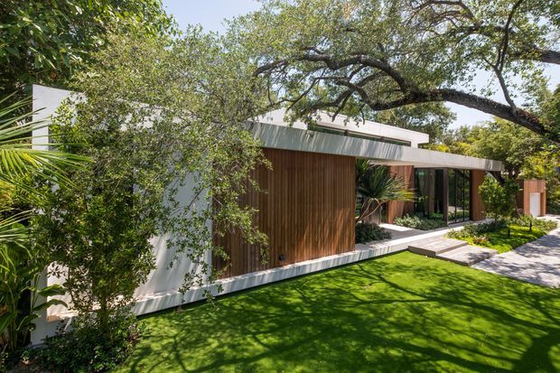 Single-storey contemporary residence viewed from the landscaped front garden, featuring crisp white rendered or panelled walls, warm vertical timber cladding, extensive dark-framed glazing, and a low flat roof with a raised clerestory-like element. The house is integrated into a mature, leafy garden with a manicured lawn and stepping-stone path.