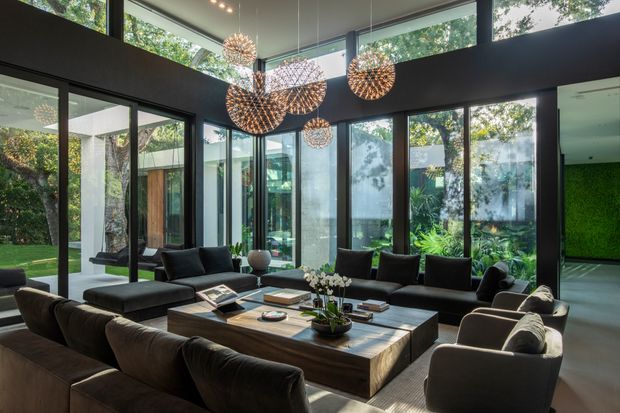 A sophisticated contemporary living room with a large sectional sofa, multiple dark upholstered lounge chairs, and a substantial timber coffee table. Floor-to-ceiling black-framed glazing wraps the room, opening views to lush greenery and an adjoining outdoor courtyard. High-level clerestory windows and a cluster of sculptural spherical pendant lights create an airy, dramatic interior with strong indoor-outdoor connection.
