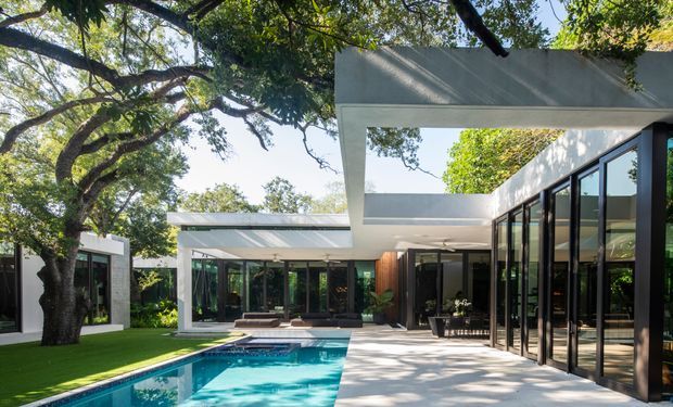 A contemporary single-storey home arranged around a swimming pool and landscaped courtyard. The design features crisp white flat-roofed volumes, extensive black-framed floor-to-ceiling glazing, open covered terraces, and strong indoor-outdoor connections beneath mature trees. Bright daylight, reflective pool water, and geometric architectural forms create a calm, resort-like setting.