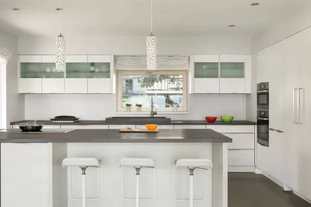 Bright contemporary kitchen with a large dark island, three white bar stools, extensive white cabinetry, integrated appliances, pale walls, and decorative white pendant lights. A window above the rear bench provides natural light and views outside.