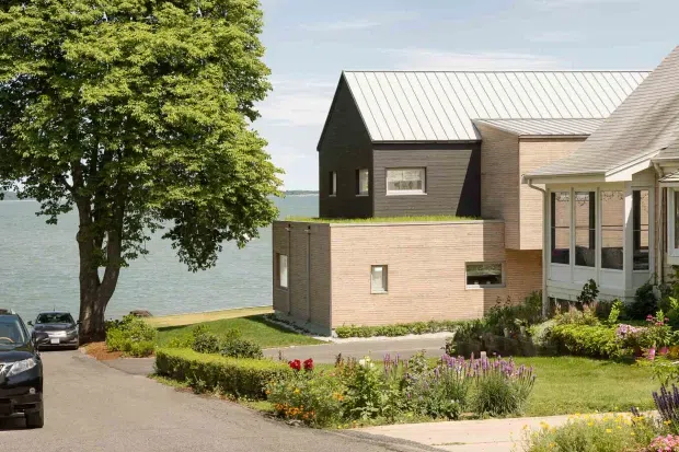 Exterior view of a contemporary multi-level residential building on a landscaped waterfront site. The composition shows pale brick and dark horizontal cladding, standing-seam metal gabled roofs, large glazed areas, a paved driveway, hedges, and garden beds, with a body of water visible behind the property.