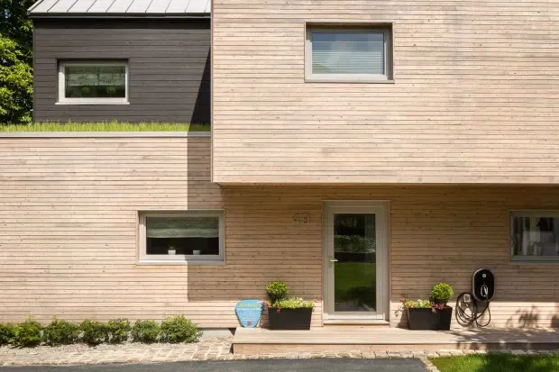 Exterior view of a contemporary multi-level house clad in horizontal light timber boards, with a projecting upper volume, dark contrasting side wall, metal roof, rectangular windows, glazed entry door, and simple planters along the paved frontage.
