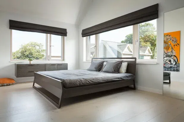 A bright contemporary bedroom with a low-profile dark timber bed, grey bedding and cushions, and matching dark roller blinds. Large corner windows provide abundant natural light and views of neighbouring homes and trees. A low timber dresser sits beneath the left window, while a colourful artwork adds a small accent to the otherwise restrained neutral scheme. The room has pale wide-plank flooring and white walls.