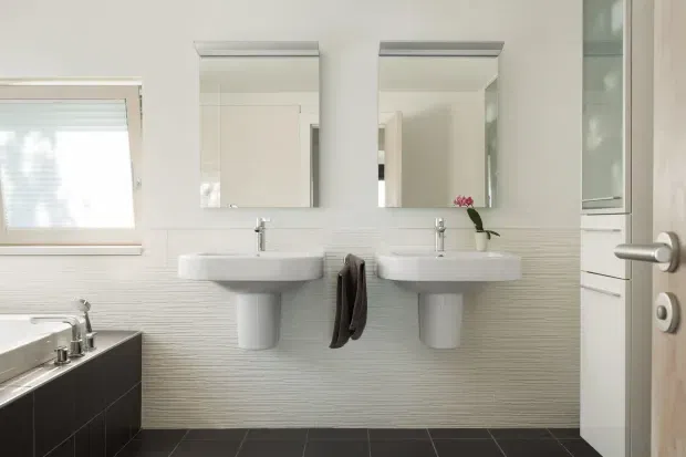 Bright contemporary bathroom with two wall-mounted white pedestal basins, matching rectangular mirrors, chrome tapware, a textured horizontal wall finish, dark tiled flooring, and dark cabinetry at the left. A window provides natural light, while an open door and partial adjacent cabinetry are visible at the right.