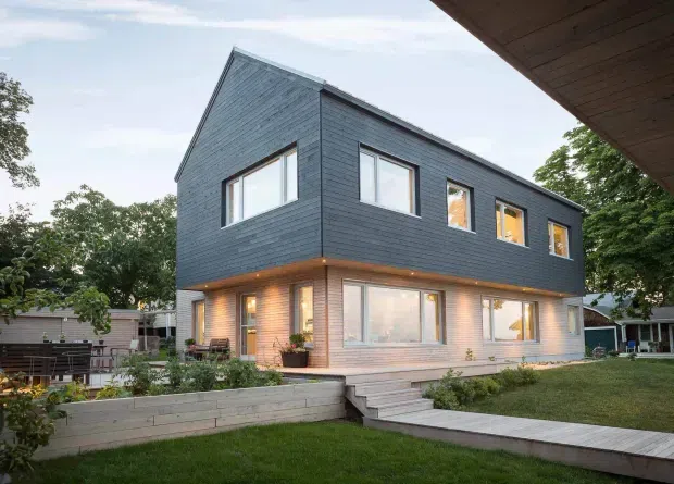 Exterior view of a contemporary two-storey residential house with a sharply pitched gable-like roof form, dark horizontal cladding on the upper level, lighter timber-toned cladding at ground level, and extensive rectangular windows. A raised timber deck and steps connect the house to a landscaped lawn, with low retaining walls and established suburban vegetation.