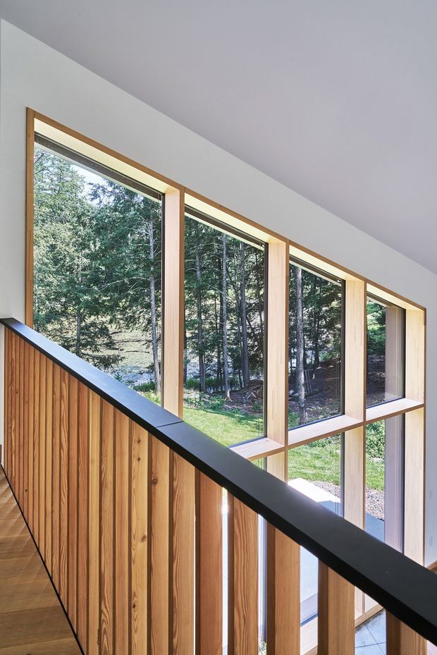 Interior view of a modern stairway or landing overlooking a tall wall of floor-to-ceiling windows. The staircase has vertical timber balustrade cladding and a slim black handrail, while the window frames and surrounding reveals are pale timber. A wooded landscape is visible outside, and the space is bright, minimal, and strongly connected to nature.