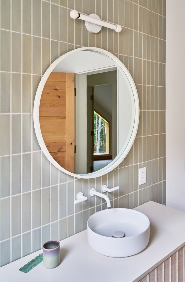 A bright, compact bathroom vanity featuring a round white vessel basin on a pale countertop, white wall-mounted tapware, and a large circular mirror. The wall behind the vanity is clad in vertically oriented muted sage-green rectangular tiles with light grout. A white globe vanity light sits above the mirror, and the mirror reflects a timber door and a naturally lit adjoining area. The overall design is clean, contemporary, and softly coloured.