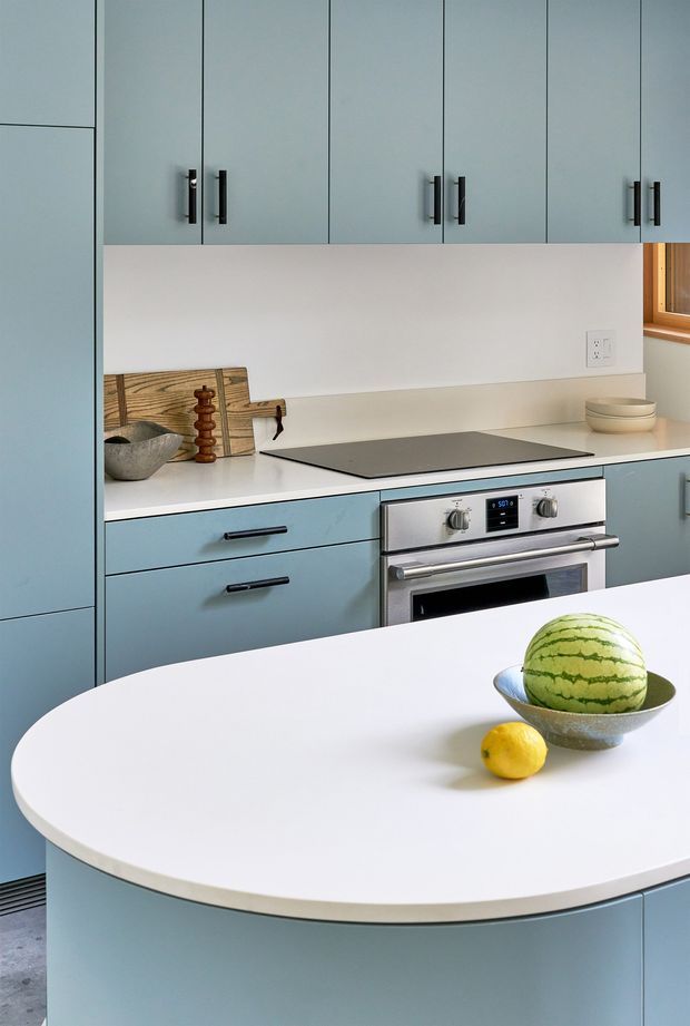 A bright contemporary kitchen with pale blue-grey flat-panel cabinetry, white benchtops, integrated upper storage, and a stainless-steel built-in oven with induction cooktop. A rounded peninsula or island with a white surface dominates the foreground, while a small timber-framed window, simple backsplash, and restrained countertop accessories complete the clean composition.
