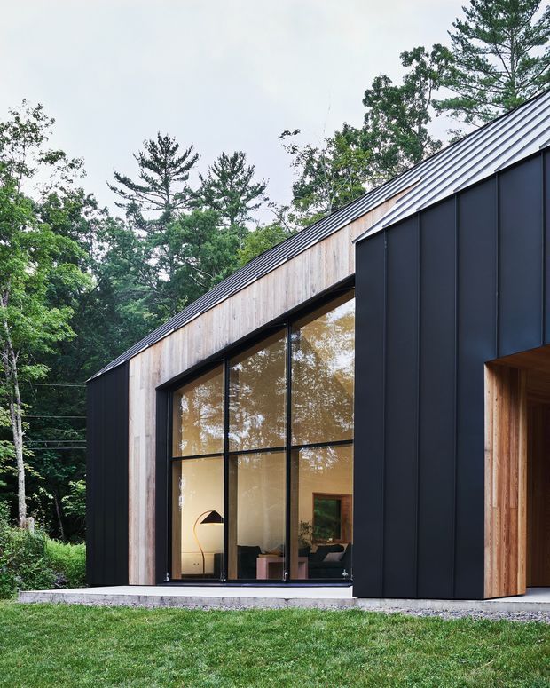 Exterior view of a contemporary single-storey building set among mature trees. The structure has a steep gabled metal roof, dark vertical metal cladding, warm timber weatherboard-style wall sections, and a large floor-to-ceiling glazed opening revealing a furnished interior. A recessed timber-lined entry is visible at right, with lawn and woodland surrounding the building.