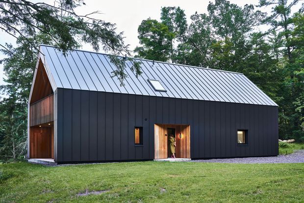 Exterior view of a compact, modern barn-like house set in a wooded rural landscape. The building has dark vertical metal cladding, a matching standing-seam pitched roof with a small roof window, warm timber-lined recessed entry and an open covered end bay. It sits on a simple gravel-edged lawn surrounded by mature trees.