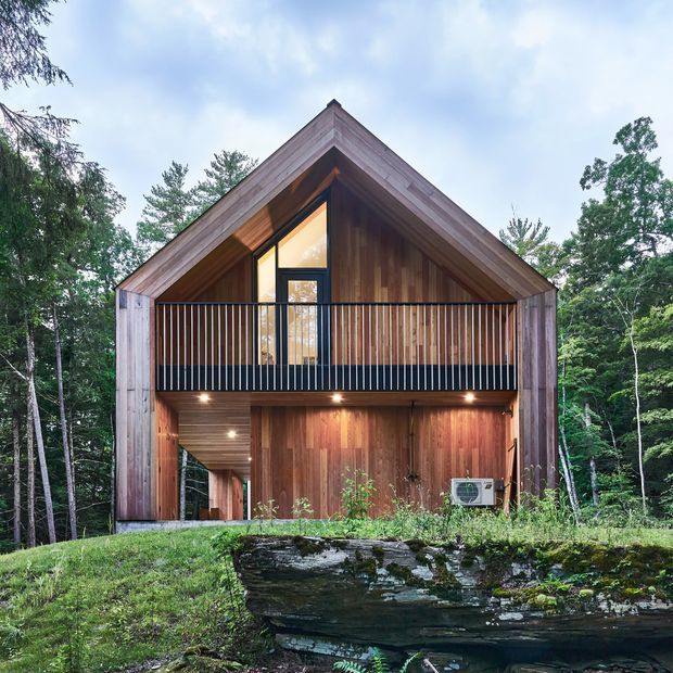 Front exterior view of a compact contemporary cabin set within a wooded landscape. The building has a steep gabled roof, extensive vertical timber cladding and detailing, a recessed upper balcony with slim black metal balustrading, and glazed openings beneath the roof peak. Warm recessed lighting illuminates the timber-lined lower level and covered outdoor area. A natural rock ledge and dense trees form the foreground and backdrop.