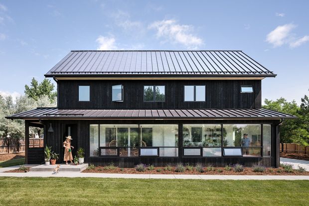 A two-storey contemporary residential house with a dark timber-clad upper level, broad metal gabled roof, and a glazed ground-floor living area extending across the front. The building has a long, low horizontal composition, dark metal roof and frames, large windows, and landscaped lawn in front.