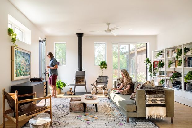 Bright, airy contemporary living room with white walls, large windows and glazed doors, a central black wood-burning stove, built-in open shelving filled with books and plants, a sofa with a patterned throw, lounge chairs, and layered rugs around a low coffee table. The room has a relaxed, organic and eclectic character with abundant natural light and greenery.