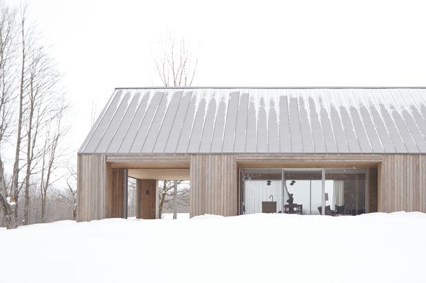 Snow-covered single-storey modern cabin or small house with a simple pitched standing-seam metal roof and vertical timber cladding. Large glazed openings reveal a sparsely furnished interior, while leafless trees and deep snow emphasize a cold rural or woodland setting.