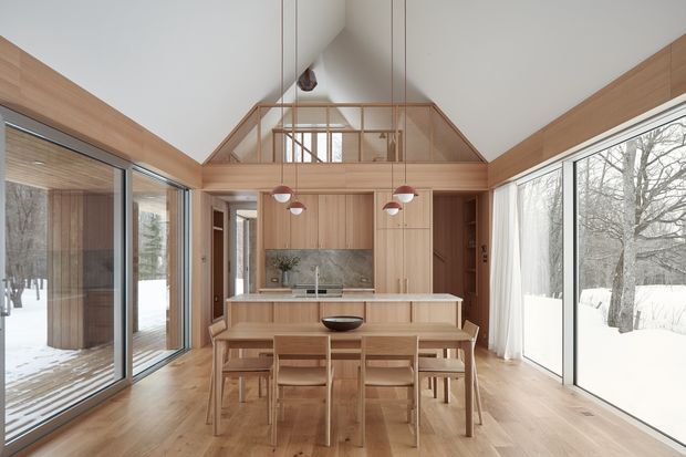 A bright, open-plan kitchen and dining area in a timber-lined gable-roofed interior. The room features extensive floor-to-ceiling glazing on both sides, pale timber flooring and wall panelling, a central island with a light benchtop, integrated timber cabinetry, a stone-look splashback, and a timber dining table with matching chairs. Snow-covered landscape and mature trees are visible outside. A glazed upper-level loft or clerestory area sits beneath the pitched roof, adding vertical openness.