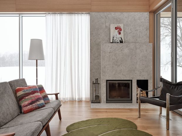 A refined contemporary living room with floor-to-ceiling glazing, pale timber wall and ceiling panelling, light timber flooring, and a textured stone fireplace surround with a built-in firebox. A grey sofa, black leather lounge chair, floor lamp, sculptural green rug, and colourful patterned cushion furnish the room. Snowy trees are visible outside through the windows.