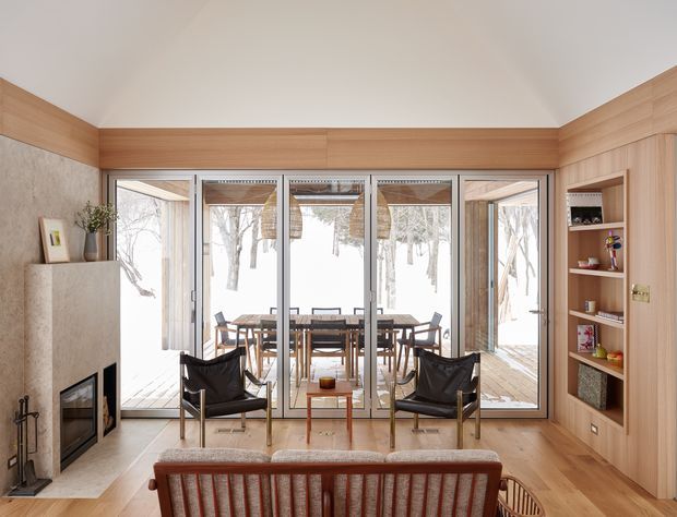 Warm contemporary living room with a built-in fireplace and timber-lined ceiling and walls, opening through full-height glazed doors to a dining area and snowy landscape beyond. The room includes a mid-century-style upholstered sofa, leather sling chairs, timber flooring, and integrated open shelving and cabinetry.