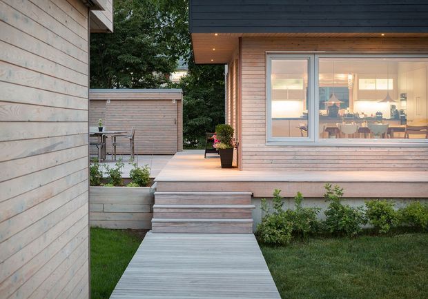 Exterior view of a contemporary timber-clad house with a light timber deck, broad steps, and a connecting boardwalk. Large windows reveal a warmly lit kitchen and dining area inside. The surrounding landscaped garden includes lawn, planted borders, and a small outdoor dining setting enclosed by timber screening.