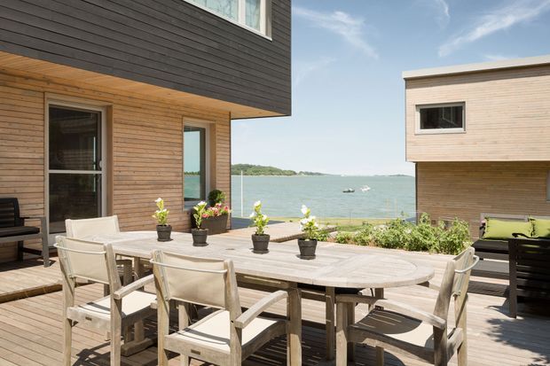 A spacious timber-clad outdoor dining terrace with a large weathered-wood dining table, surrounding armchairs, potted plants, and lounge seating. Contemporary waterfront buildings frame views across the water, creating a bright coastal setting.