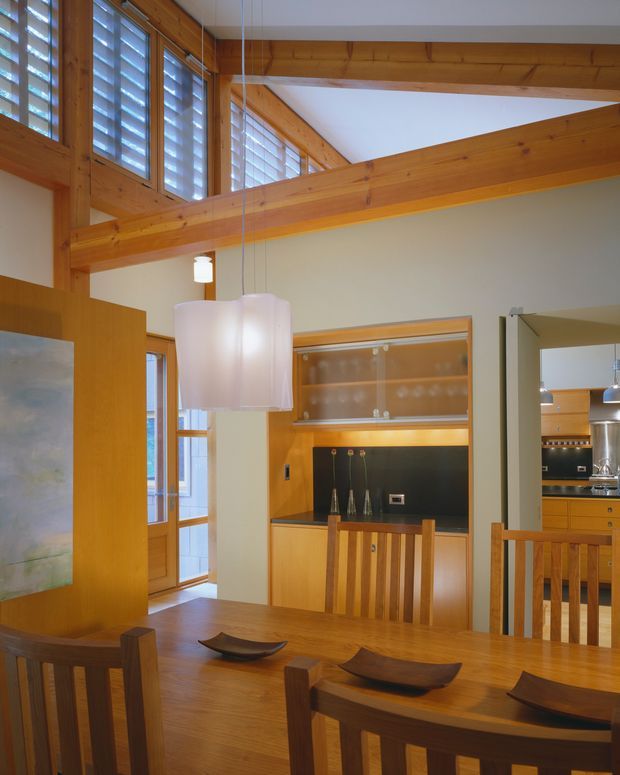 Warm timber-lined dining area with a large solid wood dining table and matching chairs, opening into a compact kitchen. Built-in timber cabinetry includes a glass-front display cabinet and dark feature backsplash. The space has exposed timber beams, high clerestory windows with louvred shutters, and pendant lighting beneath a raised ceiling.