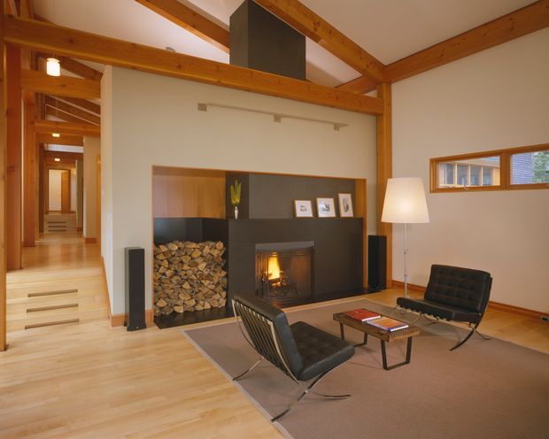 Warm contemporary living room with a built-in fireplace and firewood storage, exposed timber beams, pale timber flooring, and a hallway leading to stepped circulation at left. Black lounge chairs and a low coffee table sit on a large neutral rug, while a narrow horizontal window brings in daylight.